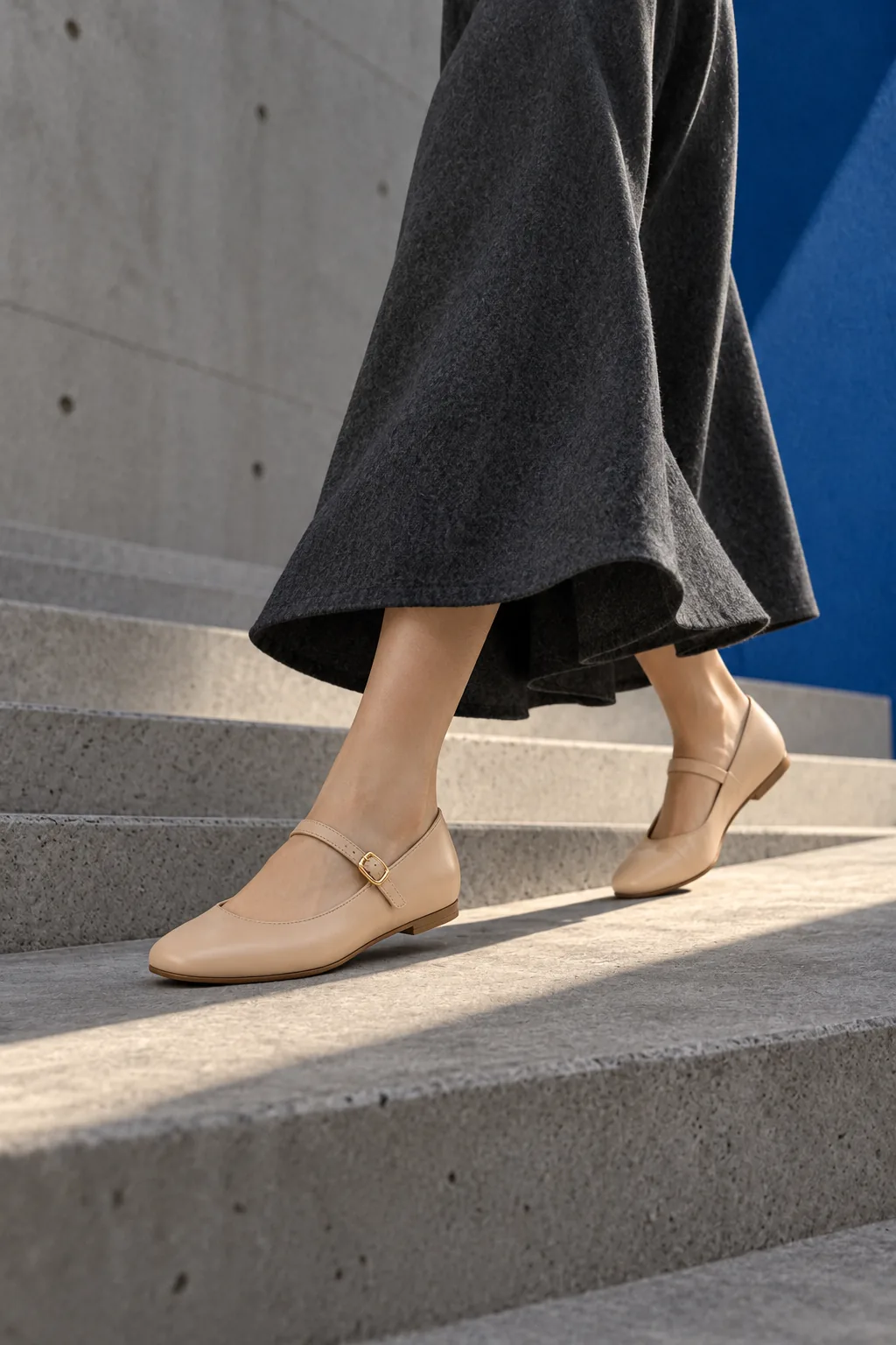 Nude leather Mary Jane flats mid-stride on a concrete stair beside a cobalt wall